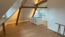 The image shows an attic or loft space with exposed wooden beams and trusses. The floor is made of hardwood planks, and there is a kitchen area visible in the background. The space has a cozy and rustic feel with the wooden elements.