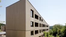 Modern wooden building, multiple windows, balconies, surrounded by greenery