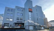 A multi-story commercial building with a gray metal facade and large windows. The building has the name 'Klubus' displayed prominently on the front. There are parking spaces in front of the building.