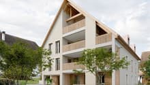 3-story apartment building with a modern, minimalist design. The building features a mix of concrete and wood materials, with large balconies on each floor. The building is surrounded by greenery and has a paved walkway in the front.