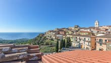 Picturesque coastal town with colorful buildings, red tile roofs, and a church steeple overlooking the sea. The town is situated on a hillside with lush greenery surrounding it.