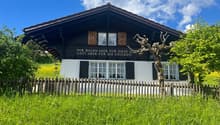2 story chalet house with brown roof, white walls, wooden shutters, and wooden fence in front, surrounded by grass