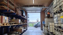 Warehouse with wooden shelves, metal racks, and storage boxes. Open door leading to a road with a van parked outside.