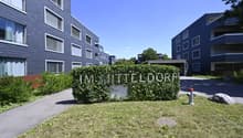 Entrance to a residential building complex with a sign reading 'IM MITTELDORF', surrounded by greenery and a pathway.