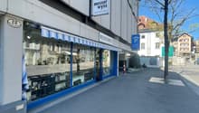 Commercial building with a shopfront, blue awning, and a sign reading 'Messer Wyss'. The building has a modern design with glass windows and a white wall. There is a parking sign and a tree in front of the building.
