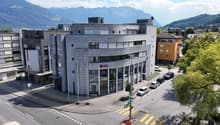 Large modern office building with concrete exterior, multiple floors, and parking spaces in front of the building. The building has the UBS logo displayed on the facade, indicating it is likely an office for the UBS financial services company. The surroun
