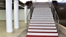 The image shows a set of stairs with a red carpet leading up to the second floor. The stairs are made of granite or a similar stone material, with metal railings on both sides. The stairs are located in a large open space with white columns supporting the