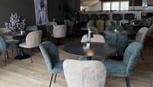 Modern indoor dining area with round tables and chairs, wooden floor, round lamps on tables, decorative plants, bar counter with chairs, and artwork on the wall.