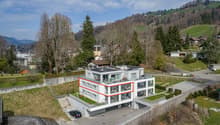 Modern white building, multiple floors, solar panels on the roof, balconies, parking area, surrounded by greenery