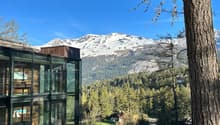 A glass-enclosed building with a view of snow-capped mountains and a forested valley in the background. The building appears to be situated in a scenic, mountainous area.