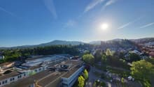 The image shows an aerial view of a town or city surrounded by mountains and forests. The town has a mix of residential and commercial buildings, with a river or stream running through the center. There are cars parked on the streets and trees lining the