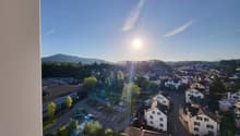 The image shows a panoramic view of a small town or city surrounded by hills and mountains. The town has a mix of residential and commercial buildings, with trees and greenery visible throughout. The sky is clear and blue, with the sun shining brightly. T