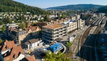 Aerial view of a cityscape with various buildings, trees, and a railway station in the foreground. The buildings are a mix of architectural styles, with some having red roofs. The trees are lush and green, and the railway station has several tracks and a
