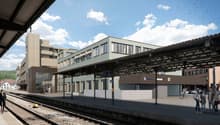 The image shows a modern train station with a covered platform and several buildings surrounding it. The station has a large, open design with glass walls and a metal roof structure. There are people waiting on the platform, and birds can be seen flying o