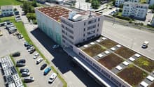 Large office building with green roof, cars parked in front of the building, adjacent parking lot.