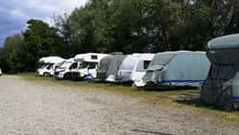 Multiple covered caravans parked in a gravel parking lot with trees in the background.
