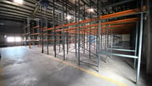 Empty warehouse with metal shelving units, concrete floor, yellow line on the floor, windows on the left side, metal beams on the ceiling