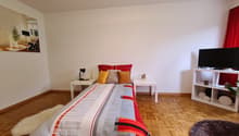 Spacious bedroom with hardwood flooring, white walls, a large bed with colorful bedding, side tables, a lamp, and a TV stand. The room also has a window, suggesting the presence of a balcony or terrace.