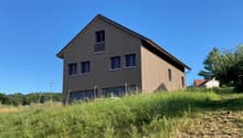 2-story wooden house with dark siding, large windows, and a balcony on the upper level. The house is situated on a grassy hill with trees in the background.