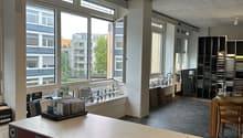 An empty office space with large windows, large sink, kitchen island, wooden table and chairs, kitchen cabinets, and racks with various items. There is a large glass window, with a view of the buildings outside. The room is mostly empty, with several furn