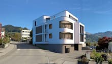 Modern white building, multiple balconies, large windows, parking area, mountains in the background