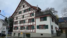 White painted, timber-framed house, red roof, multiple windows, red trims, small garden