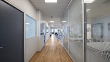 Office hallway with wooden flooring, glass partitions, and overhead lighting.