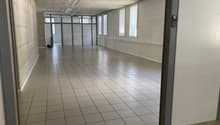 An empty, rectangular room with white walls, tiled flooring, and windows with blinds. The room is illuminated by white ceiling lights. It appears to be vacant with no furniture or decorations.