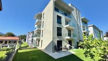A three-story apartment building with modern design, balconies, and large windows, surrounded by a well-maintained lawn and lush greenery, creating a peaceful and picturesque environment.