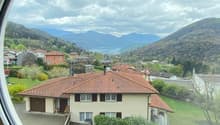 The image shows a scenic view through a round window. It depicts a mountainous landscape with a small town nestled in the valley below. The town has residential buildings with red tile roofs, surrounded by lush greenery and trees. In the distance, there a