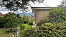 2-story house surrounded by lush greenery, including trees and shrubs. The house has a balcony or terrace overlooking the scenic mountain landscape in the background.