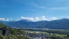 The image shows a panoramic view of a mountainous landscape with a town or village nestled in the valley below. The mountains in the background are tall and rugged, with a blue sky dotted with clouds overhead. The foreground includes some buildings or str