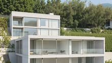 Modern two-story house with large glass windows, balconies, and surrounded by lush greenery