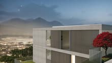 Modern concrete and glass building with balconies, situated on a hillside overlooking a city with mountains in the background. The building has a minimalist, geometric design with horizontal and vertical lines. There is a red tree in the foreground, addin