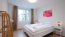 Spacious bedroom with white walls, large windows, hardwood flooring, a white bed with white bedding, two bedside tables with lamps, and a colorful abstract painting on the wall. The room also has a wooden ladder-like structure, possibly for storage or dec