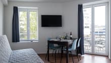 The image shows a studio apartment with a living/sleeping area, a dining area, and a balcony. The living area has a large window with a view of the outdoors, a gray quilted bedspread, and a mounted TV. The dining area has a dark wooden table with blue cha