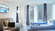 Spacious bedroom with large windows, gray sofa, laptop on a table, and a bed with blue bedding
