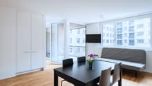 Spacious open-plan living area with large windows, hardwood floors, black dining table and chairs, gray sofa, built-in wardrobe, and access to a balcony or terrace with a view of the surrounding buildings.