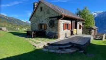 Single story stone house, red shutters, slate roof, porch, wooden wheel, located in a mountainous area with a view of the peaks