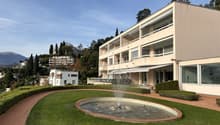 Modern apartment building with balconies, surrounded by greenery and a fountain, with a scenic view of mountains