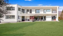 Two-story modern white house with large windows, balcony, and patio area overlooking a well-manicured lawn and garden. The house has a clean, minimalist design with a flat roof and appears to be situated in a scenic, mountainous area with a clear blue sky