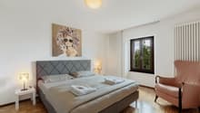 Spacious bedroom with a large bed, gray upholstered headboard, artwork on the wall, and a window overlooking the outdoors.