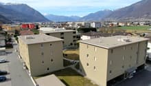 The image shows a residential area with several multi-story apartment buildings. The buildings have a beige exterior and are surrounded by paved areas and small grassy spaces. There appears to be a parking area visible in the foreground. The buildings are