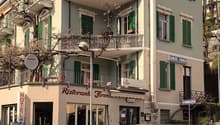 3-story building, white exterior, green shutters, balcony on first and second floor, awnings, restaurant on the ground floor, garage entrance