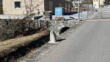 A single-story gray building with a slanted roof, surrounded by a stone wall and located in a mountainous area. There is a street with a speed limit sign visible in the foreground.