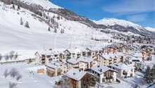 Snowy mountain landscape with a ski resort town nestled in the valley. The town features multi-story apartment buildings and chalets with balconies and parking spaces. The surrounding mountains are covered in snow, creating a picturesque winter scene.