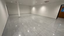 A room with white walls, ceiling, and tiled floor, and lights installed in the ceiling. There is a pillar in the middle of the room and an exit door at the right end.