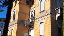 The building has multiple levels, each with balconies, a mix of beige and yellow wall colors, a tower-like section with distinct windows and arches, plants and trees around the area, and a shadow cast on the lower section.
