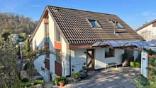 Detached house, brown roof, white walls, brown trim, two windows, two dormers, covered entrance, paved driveway, garden with plants and flowers