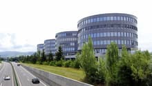 Multi-story office building with curved glass facade, surrounded by trees and a highway with cars passing by
